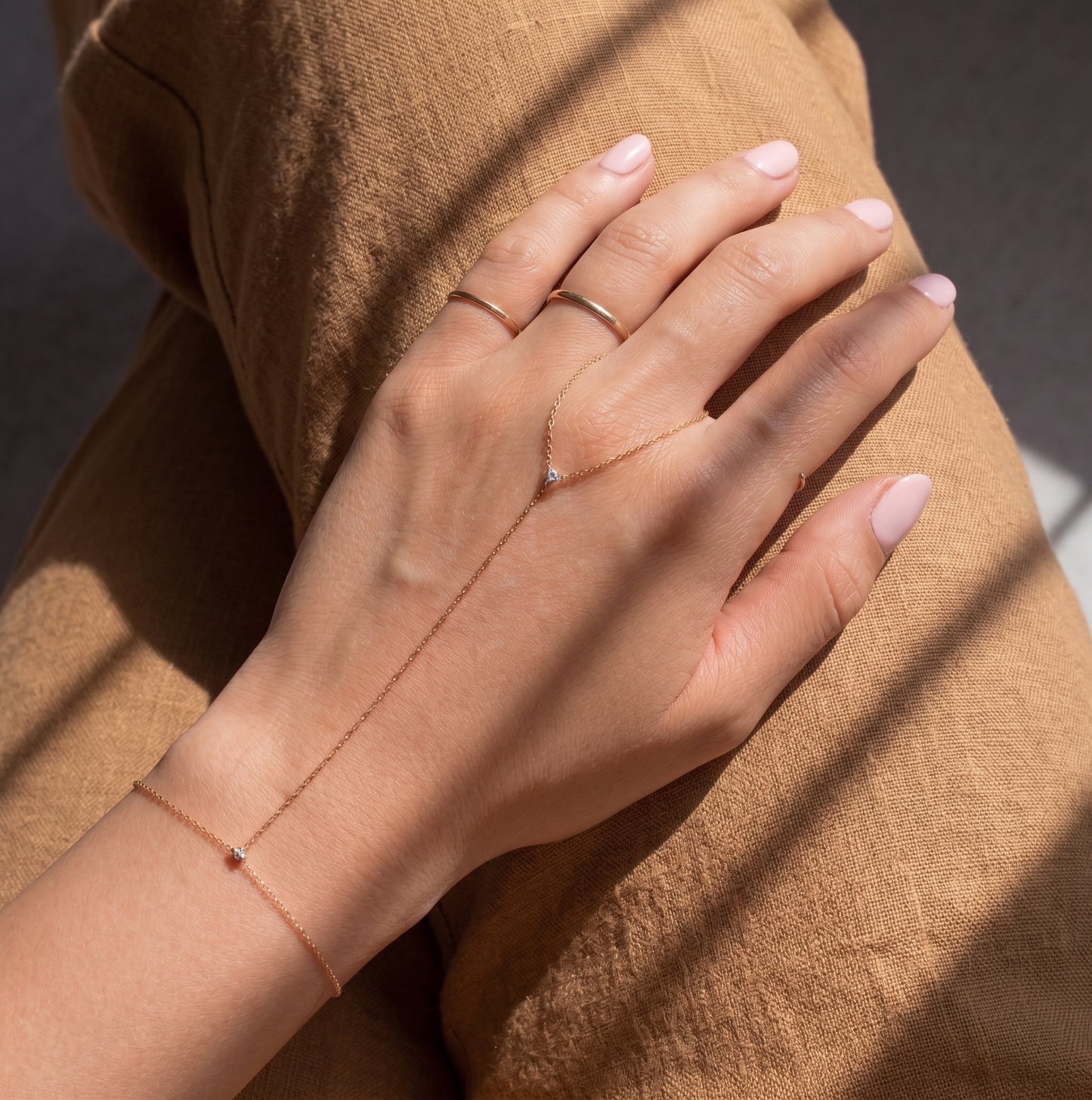 A delicate rose gold chain hand bracelet and matching ring worn on a model's hand, resting on brown linen fabric under soft window shadows.