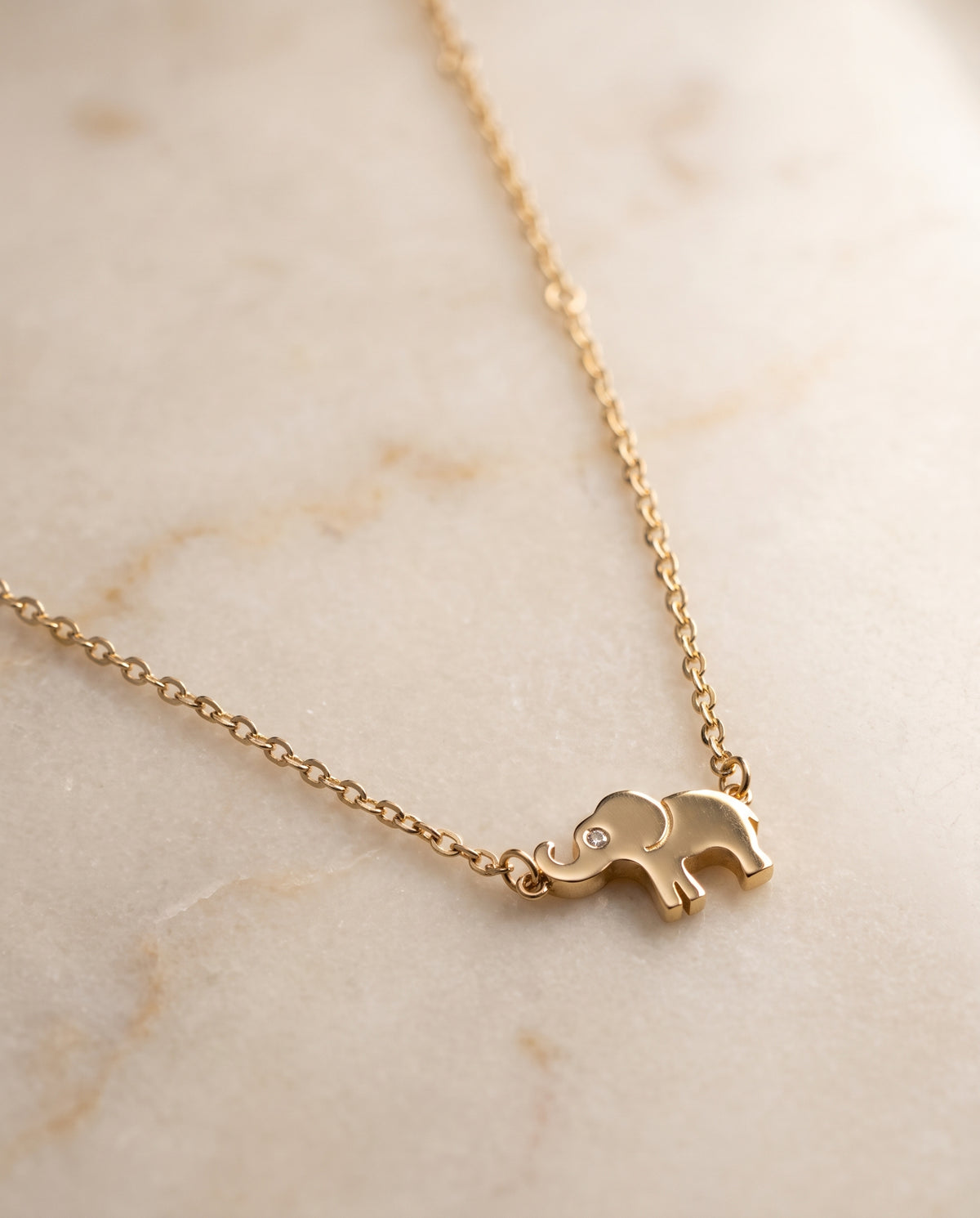 A delicate gold chain necklace featuring a small elephant pendant with a tiny clear stone in its eye, resting on a light beige marble surface.