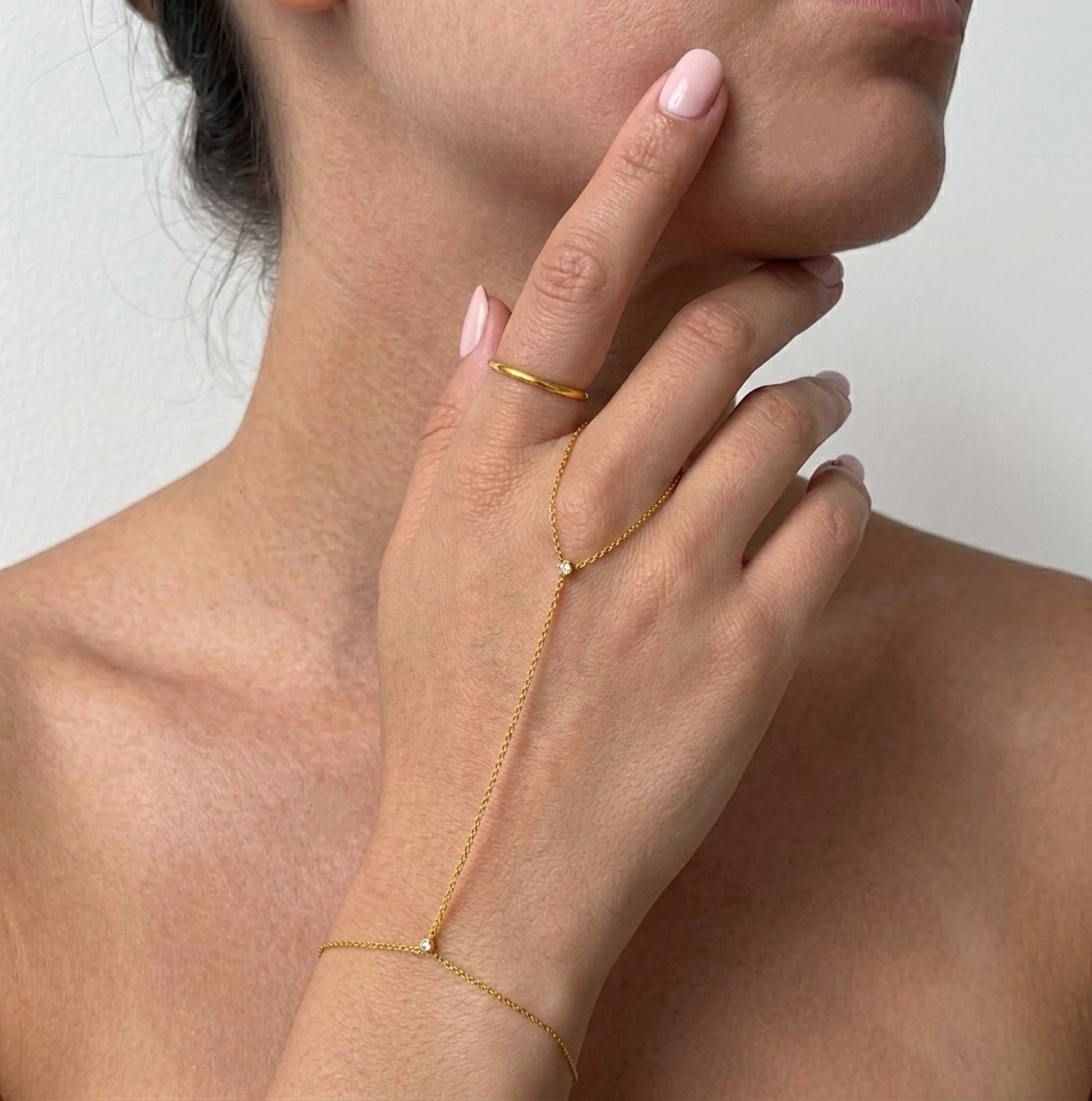 A close-up of a model's hand and neck wearing a delicate gold chain hand bracelet and a matching gold ring with small clear stones.