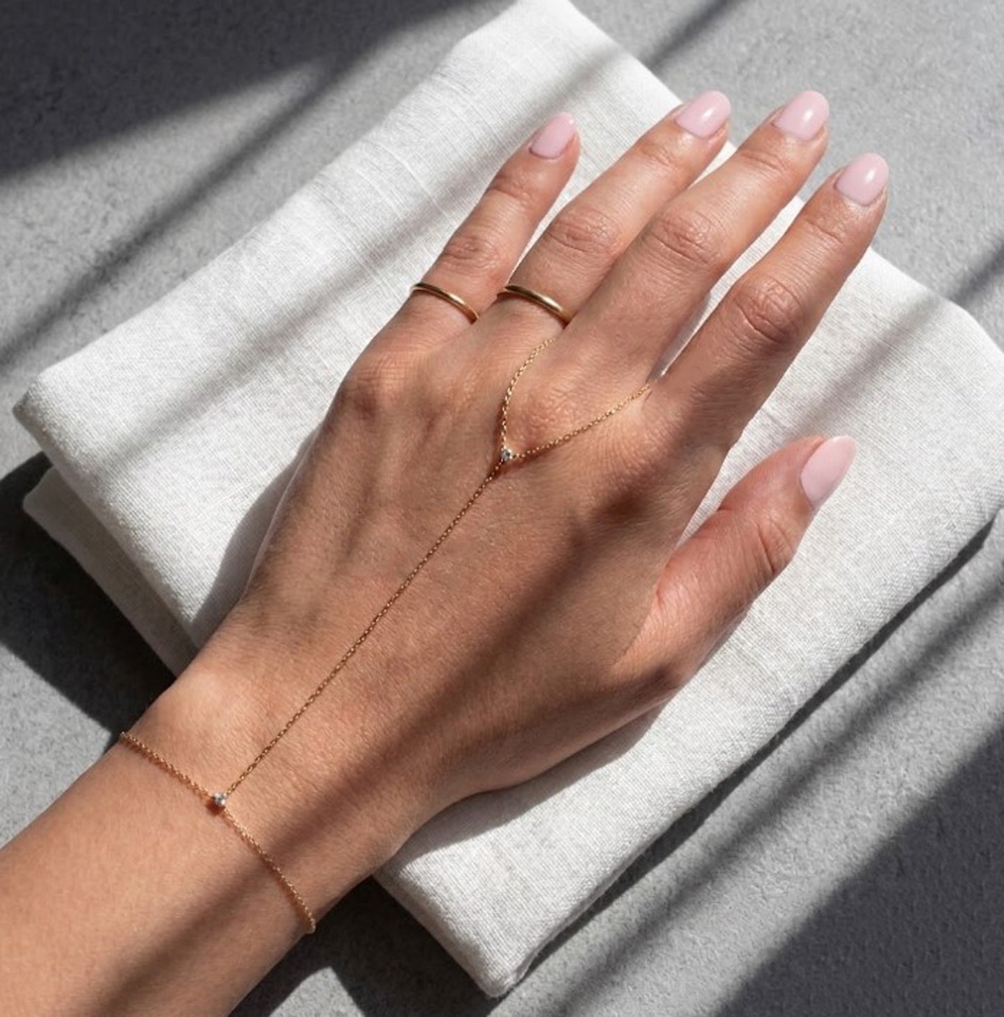 A delicate rose gold chain jewelry set worn on a model's hand and wrist, featuring a Y-shaped ring with a small clear stone and a matching bracelet, displayed on a folded white linen cloth with soft shadows.