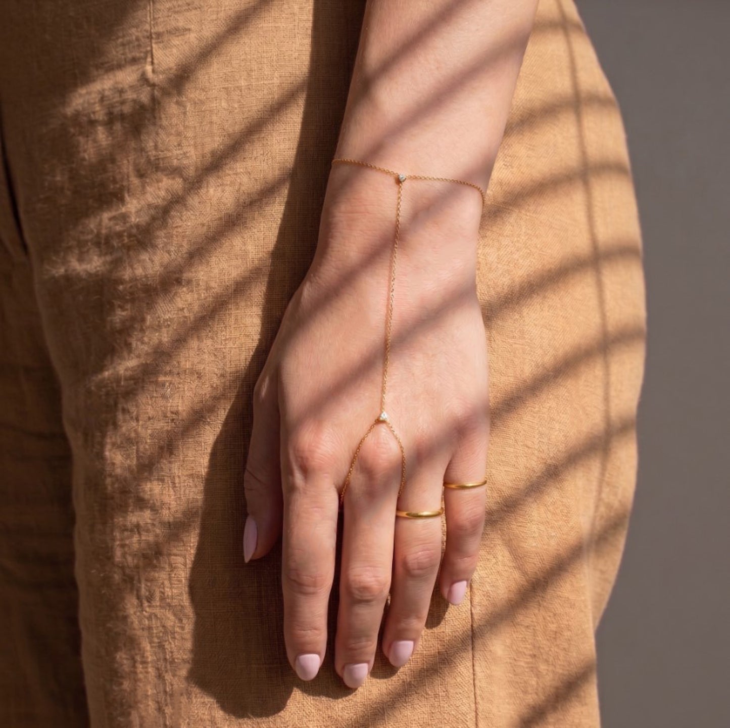 A delicate gold chain hand bracelet and matching rings worn on a model's hand, styled with a tan linen dress under soft window shadows.