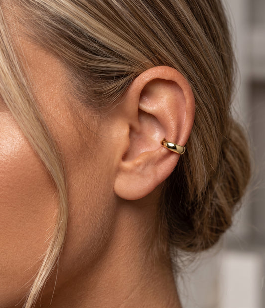 A close-up side profile of a woman's ear wearing a small polished gold hoop earring on the helix.