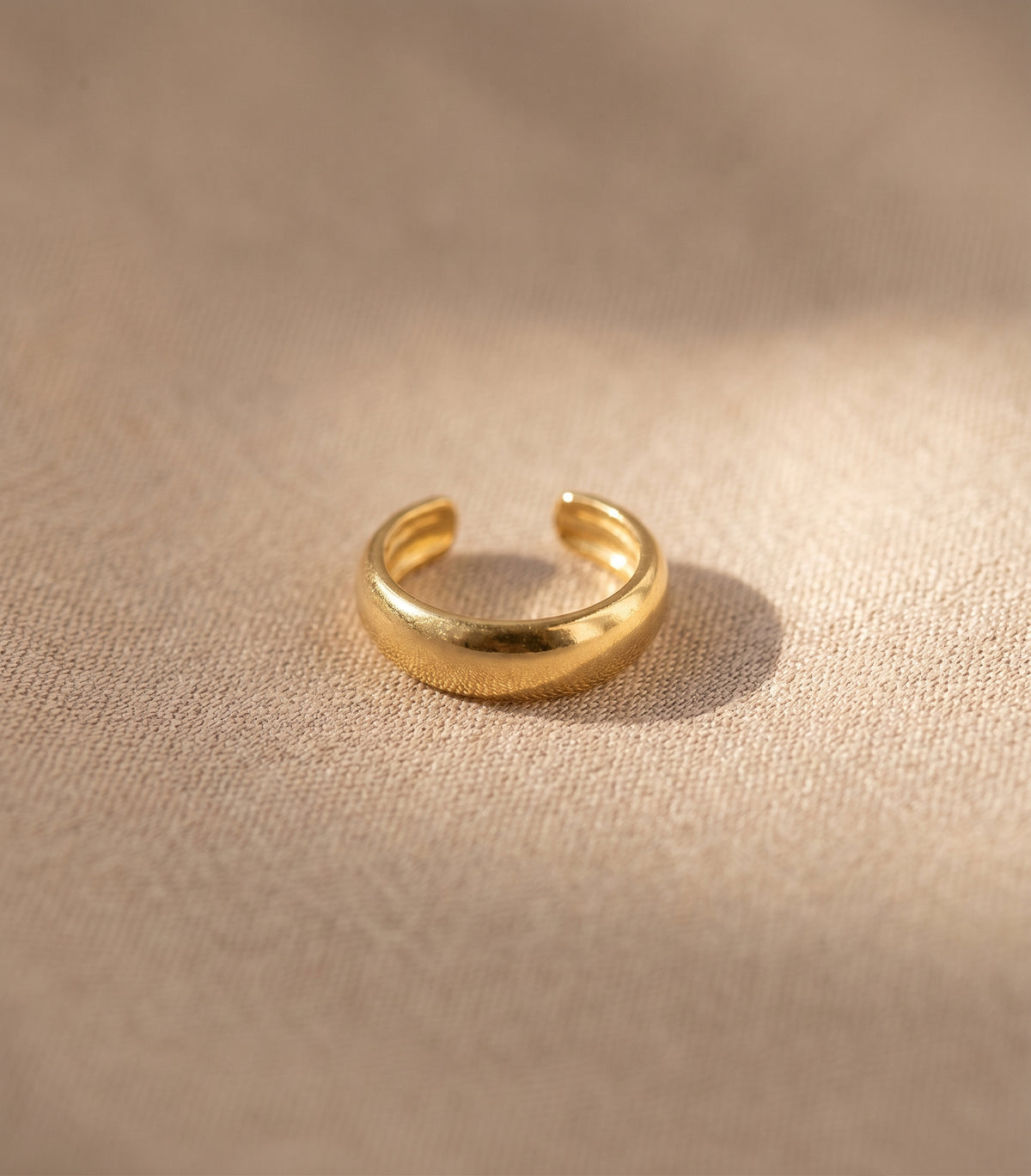 A polished gold open-ended ring, also known as a stretch band or ear cuff, resting on a textured beige linen surface with soft, warm sunlight casting a shadow.